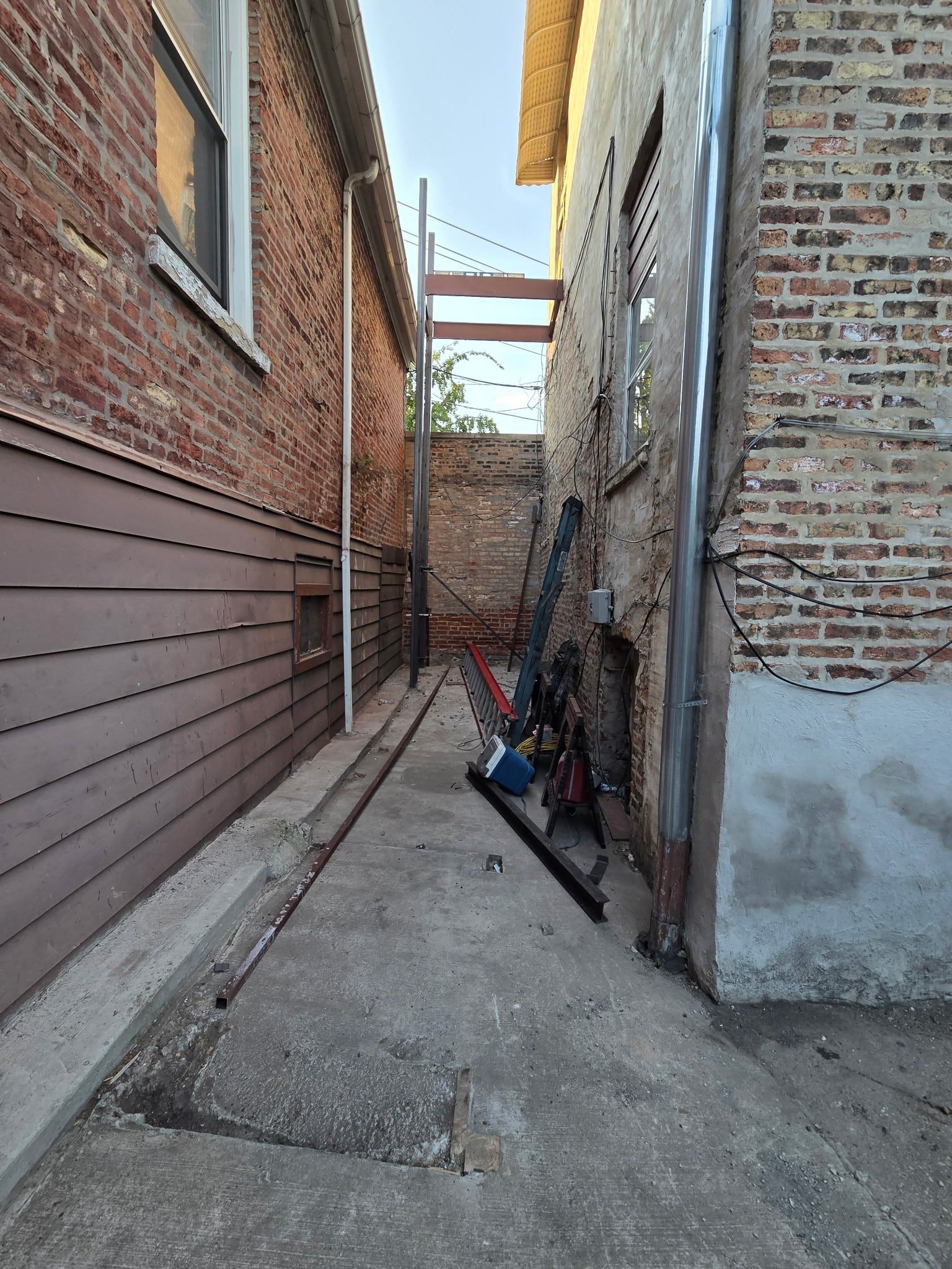 Narrow gangway between two brick buildings before the new stairs, with steel beams laid out on the ground